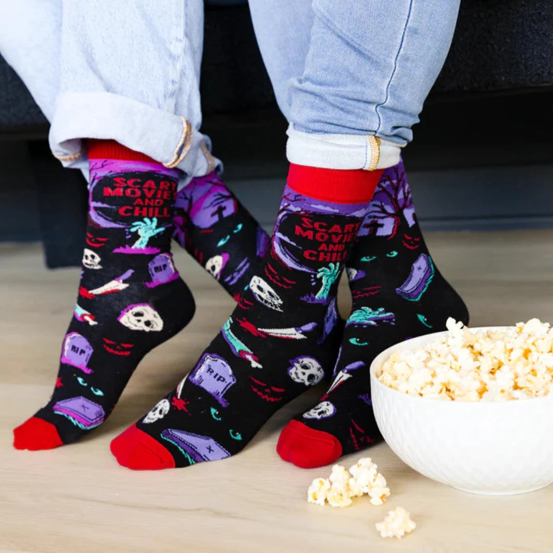A pair of crew socks with a Halloween theme. The socks are mostly black with red and purple accents. The design includes a zombie hand, a graveyard with tombstones, a skull, and bats. The text "Scary Movies and Chill" is printed on the socks.