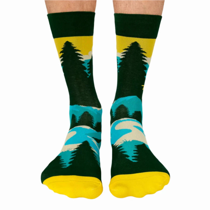 A pair of green socks with a hiking-themed design. The socks feature a mountain landscape, a setting sun, and the text 