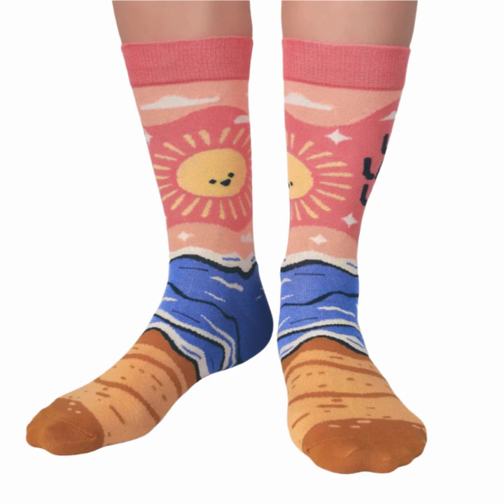 A pair of pink socks with a beach-themed design. The socks feature a beach scene, waves, and the text 