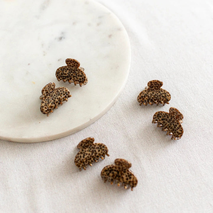 Six small, leopard print hair claw clips arranged on a white marble surface. The clips have a marbled texture.
