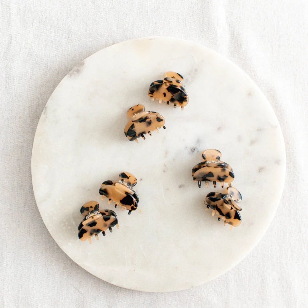 Six small, light tortoise hair claw clips arranged on a white marble surface. The clips have a marbled texture.