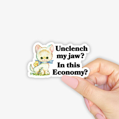 Sticker with cartoon cat and text 'Unclench my jaw? In this Economy?' held by a hand.