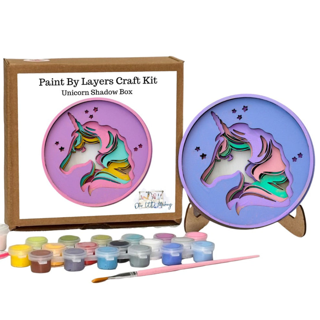 Paint by Layers Craft Kit for making a Unicorn Shadow Box, showing the kraft paper box packaging, the completed 3D layered unicorn plaque on a small wooden stand, a set of colorful acrylic paint pots, and a paintbrush on a white background.