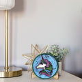 A small round decorative clock or ornament featuring a colorful unicorn illustration on a blue background, displayed on a tabletop next to a gold star decor piece and a small plant.