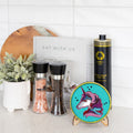 A styled kitchen countertop scene featuring a round turquoise coaster with a colorful unicorn illustration on a small wooden stand, alongside two black pepper and salt shakers, a bottle of Sparta brand olive oil, a white sign reading 'EAT WITH US', and a small potted plant against a white tiled wall.
