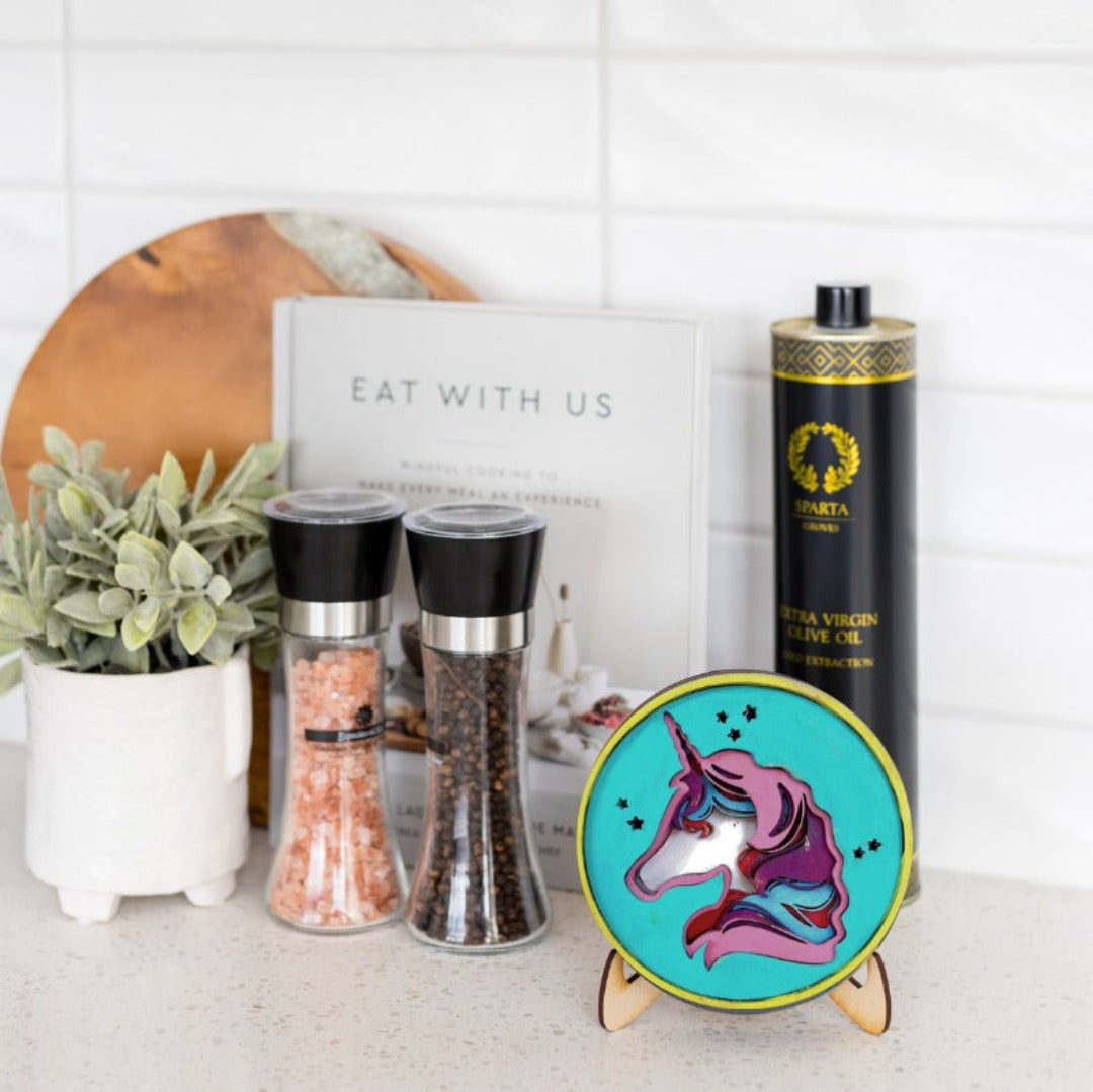 A styled kitchen countertop scene featuring a round turquoise coaster with a colorful unicorn illustration on a small wooden stand, alongside two black pepper and salt shakers, a bottle of Sparta brand olive oil, a white sign reading 'EAT WITH US', and a small potted plant against a white tiled wall.
