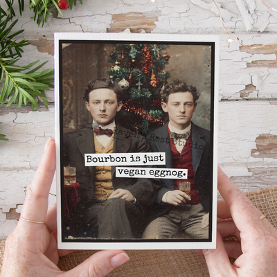 Two men holding a glass with text 'Bourbon is just vegan eggnog' against a Christmas tree background.