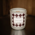 A white glass jar candle labeled 'VELVET OUD' by North Rose, featuring a geometric burgundy and white diamond pattern on the label, set on a dark wooden surface against a dark wood background.
