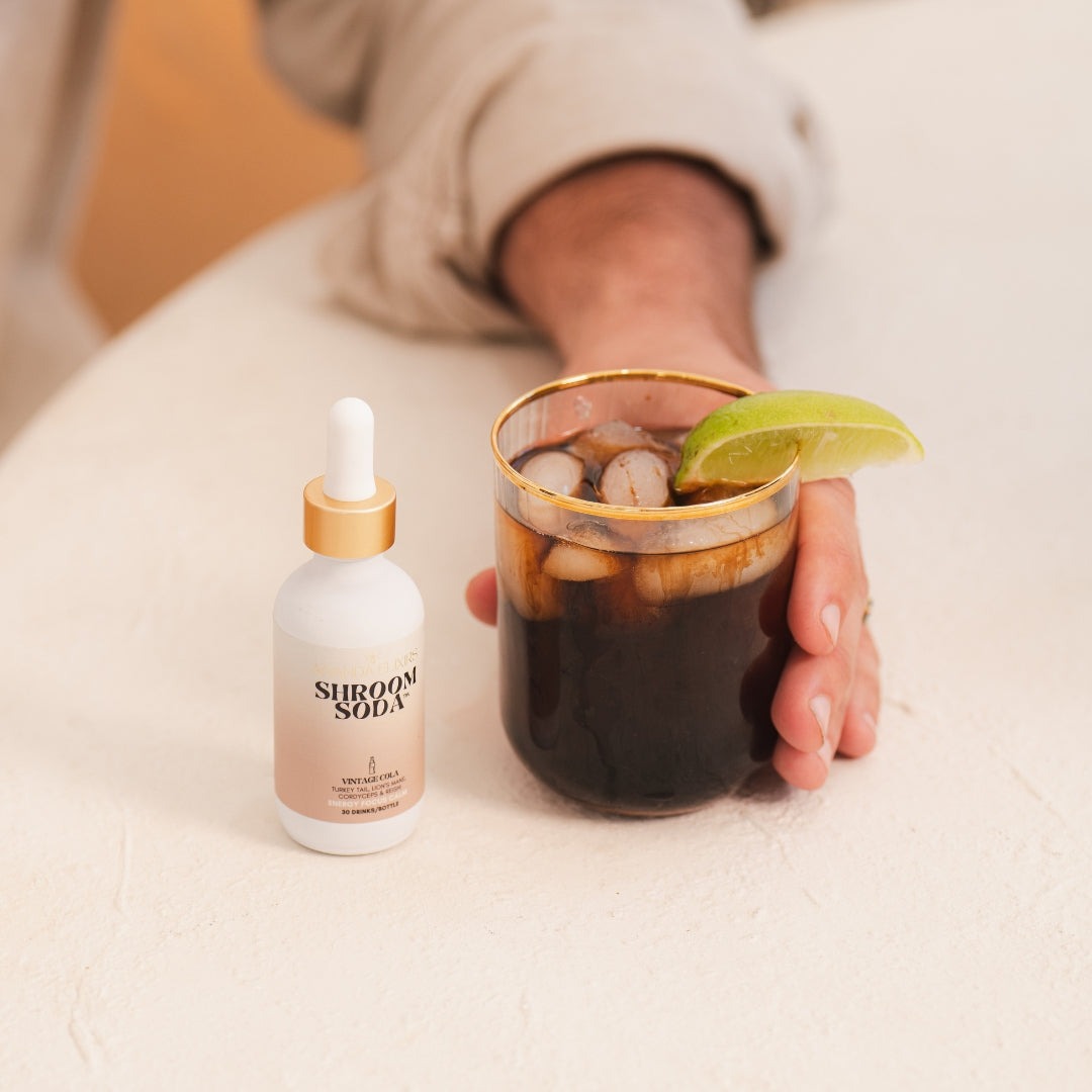 Person holding a glass of dark liquid with a lime wedge next to a bottle labeled 'Shroom Soda'.