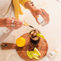 Person adding a liquid from a bottle labeled 'Wunder' to a drink on a wooden cutting board with lemon and lime slices.