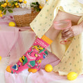 A person seated indoors wearing a bright pink novelty sock with cocktail illustrations and the word 'VODKA' on the side. The scene includes a pale yellow patterned skirt, a pink striped towel, lemons, and a pink ribbed glass.