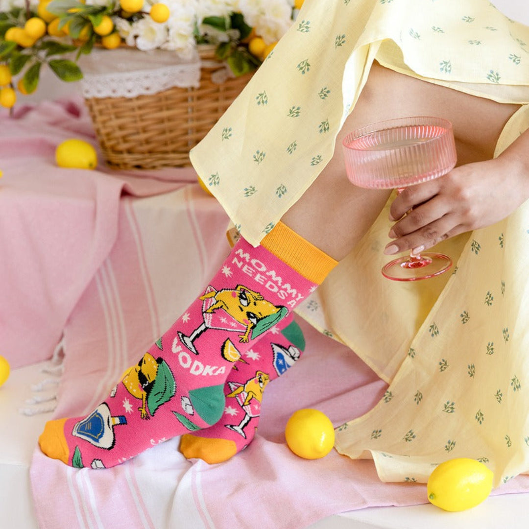 A person seated indoors wearing a bright pink novelty sock with cocktail illustrations and the word 'VODKA' on the side. The scene includes a pale yellow patterned skirt, a pink striped towel, lemons, and a pink ribbed glass.
