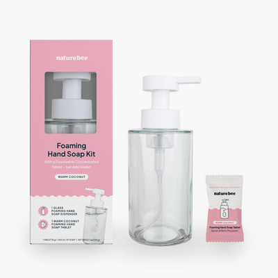 A Warm Coconut Foaming Hand Soap Kit by Nature Bee. The kit includes a pink and white box, a clear glass foaming hand soap dispenser with a white pump, and a small, individual packet of the Warm Coconut foaming hand soap tablet.
