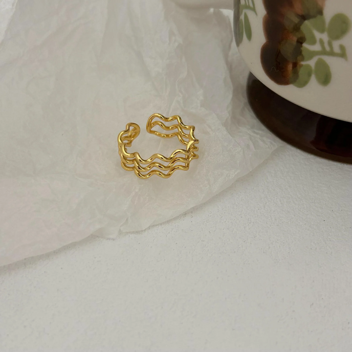 A wave ring made with 18K gold plating, featuring a modern, abstract, and undulating triple wave design, displayed on a light, textured surface. This unique ring is available in Ottawa, Ontario, Canada.