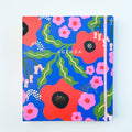 A blue agenda with a floral design. The cover features large red poppies, pink flowers, and green leaves against a blue background. The word "AGENDA" is printed in gold foil on the cover, along with the brand name "IDLEWILD CO." The agenda has a pink elastic band.