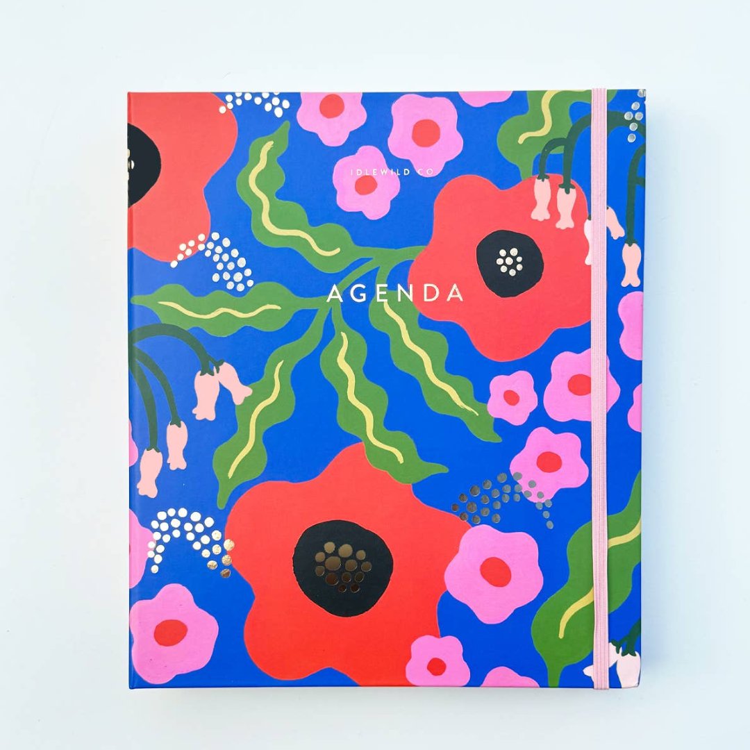 A blue agenda with a floral design. The cover features large red poppies, pink flowers, and green leaves against a blue background. The word "AGENDA" is printed in gold foil on the cover, along with the brand name "IDLEWILD CO." The agenda has a pink elastic band.