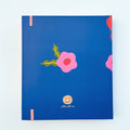A blue agenda with a floral design. The cover features large red poppies, pink flowers, and green leaves against a blue background. The word "AGENDA" is printed in gold foil on the cover, along with the brand name "IDLEWILD CO." The agenda has a pink elastic band.