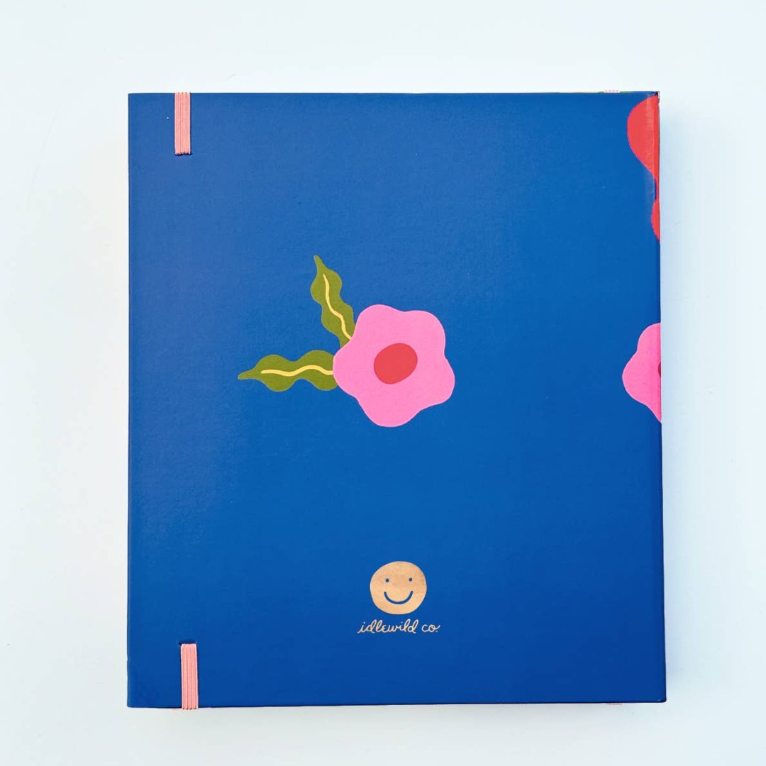 A blue agenda with a floral design. The cover features large red poppies, pink flowers, and green leaves against a blue background. The word "AGENDA" is printed in gold foil on the cover, along with the brand name "IDLEWILD CO." The agenda has a pink elastic band.