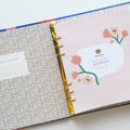 A blue agenda with a floral design. The cover features large red poppies, pink flowers, and green leaves against a blue background. The word "AGENDA" is printed in gold foil on the cover, along with the brand name "IDLEWILD CO." The agenda has a pink elastic band.