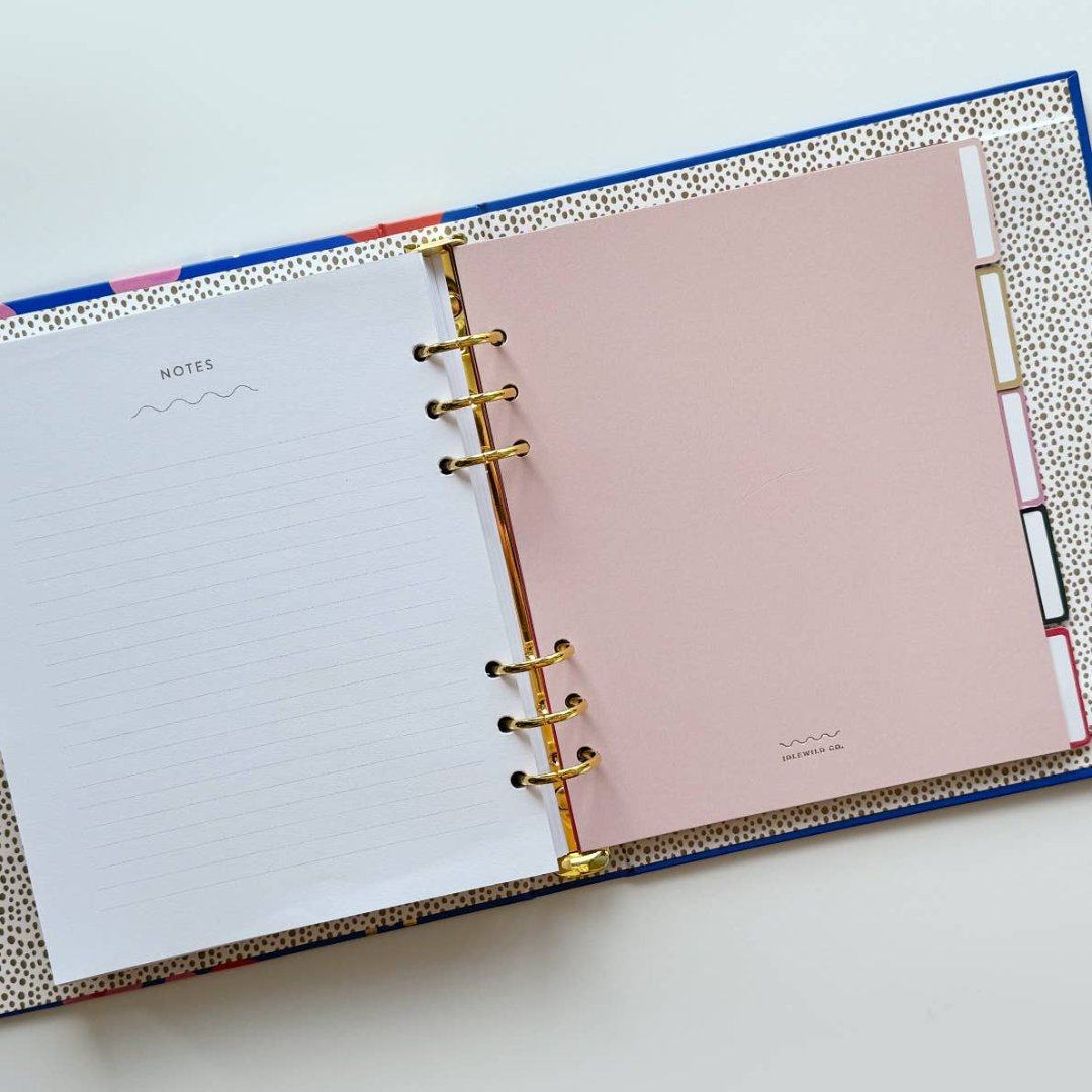 A blue agenda with a floral design. The cover features large red poppies, pink flowers, and green leaves against a blue background. The word "AGENDA" is printed in gold foil on the cover, along with the brand name "IDLEWILD CO." The agenda has a pink elastic band.