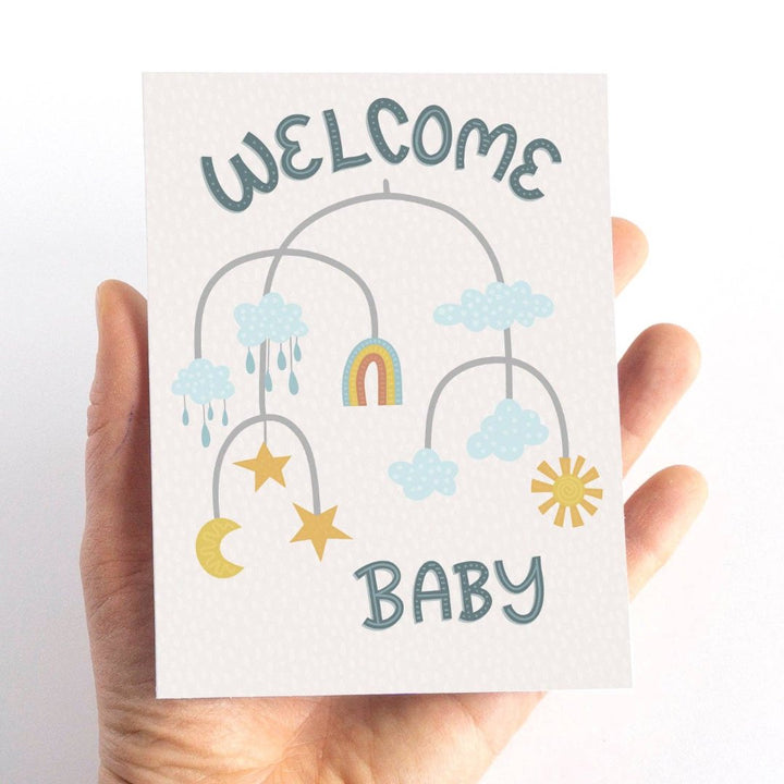 A welcome baby card featuring a mobile with clouds, a rainbow, stars, a moon, and raindrops. The words 