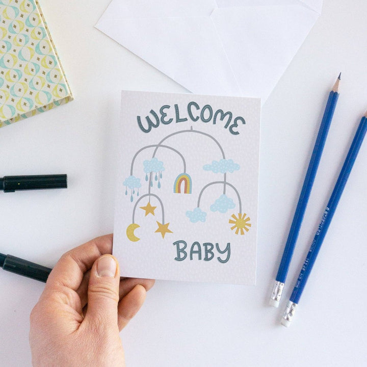 A welcome baby card featuring a mobile with clouds, a rainbow, stars, a moon, and raindrops. The words 