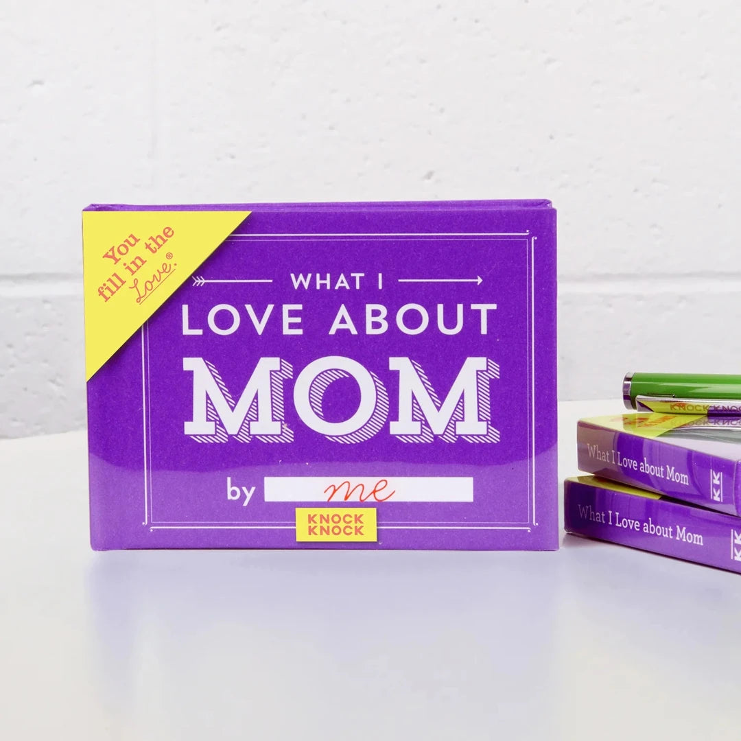 A purple book titled "What I Love About Mom" by Knock Knock. The book has a gold sticker on the front that says "YOU FILL IN THE LOVE." The cover also features the phrase "What I Love About Mom" in large, bold letters.