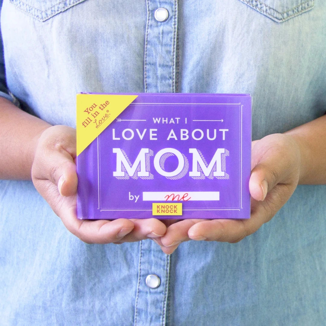 A purple book titled "What I Love About Mom" by Knock Knock. The book has a gold sticker on the front that says "YOU FILL IN THE LOVE." The cover also features the phrase "What I Love About Mom" in large, bold letters.