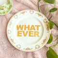 Decorative plate with 'WHAT EVER' text on a pink surface with green leaves.