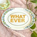 Decorative plate with 'WHAT EVER' text on a floral design, placed on a soft surface with leaves.