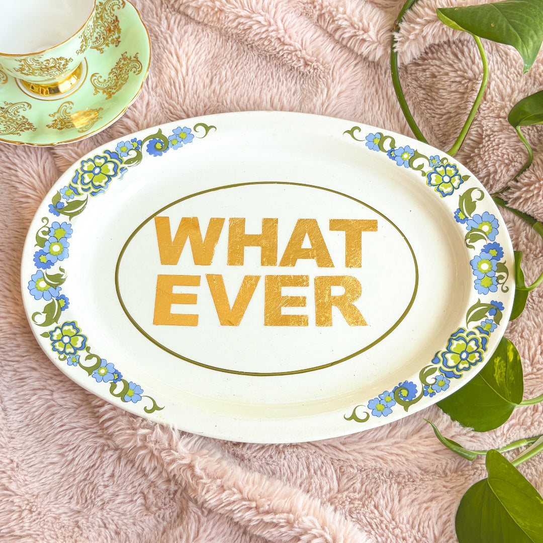 Decorative plate with 'WHAT EVER' text on a floral design, placed on a soft surface with leaves.