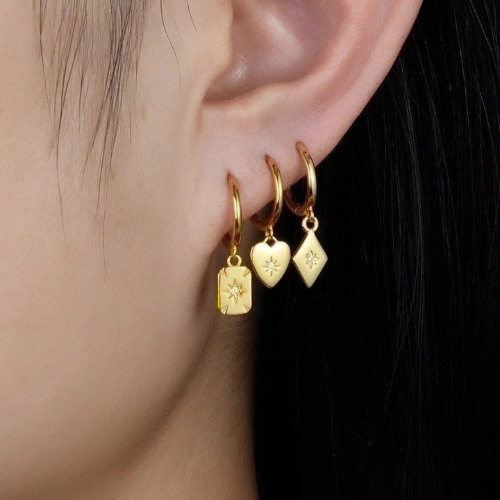 Gold plated mismatched charm hoop earrings, potentially Bohomoon style, worn on an ear with three piercings, featuring a small hoop with a rectangular charm, a heart charm, and a diamond (rhombus) charm, each with a central crystal. These unique earrings are available in Ottawa, Ontario, Canada.
