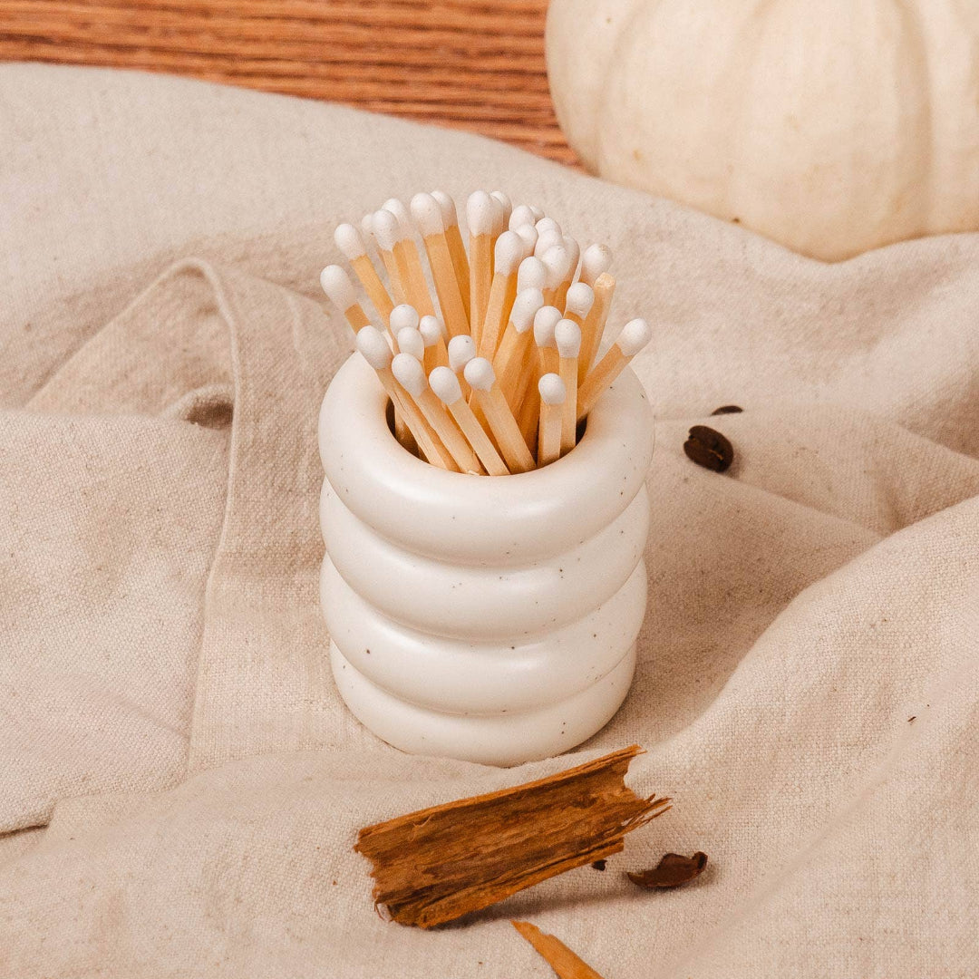 White ceramic container with matches on a beige surface with pumpkins and cinnamon sticks.