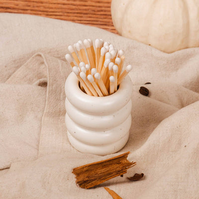 White ceramic container with matches on a beige surface with pumpkins and cinnamon sticks.