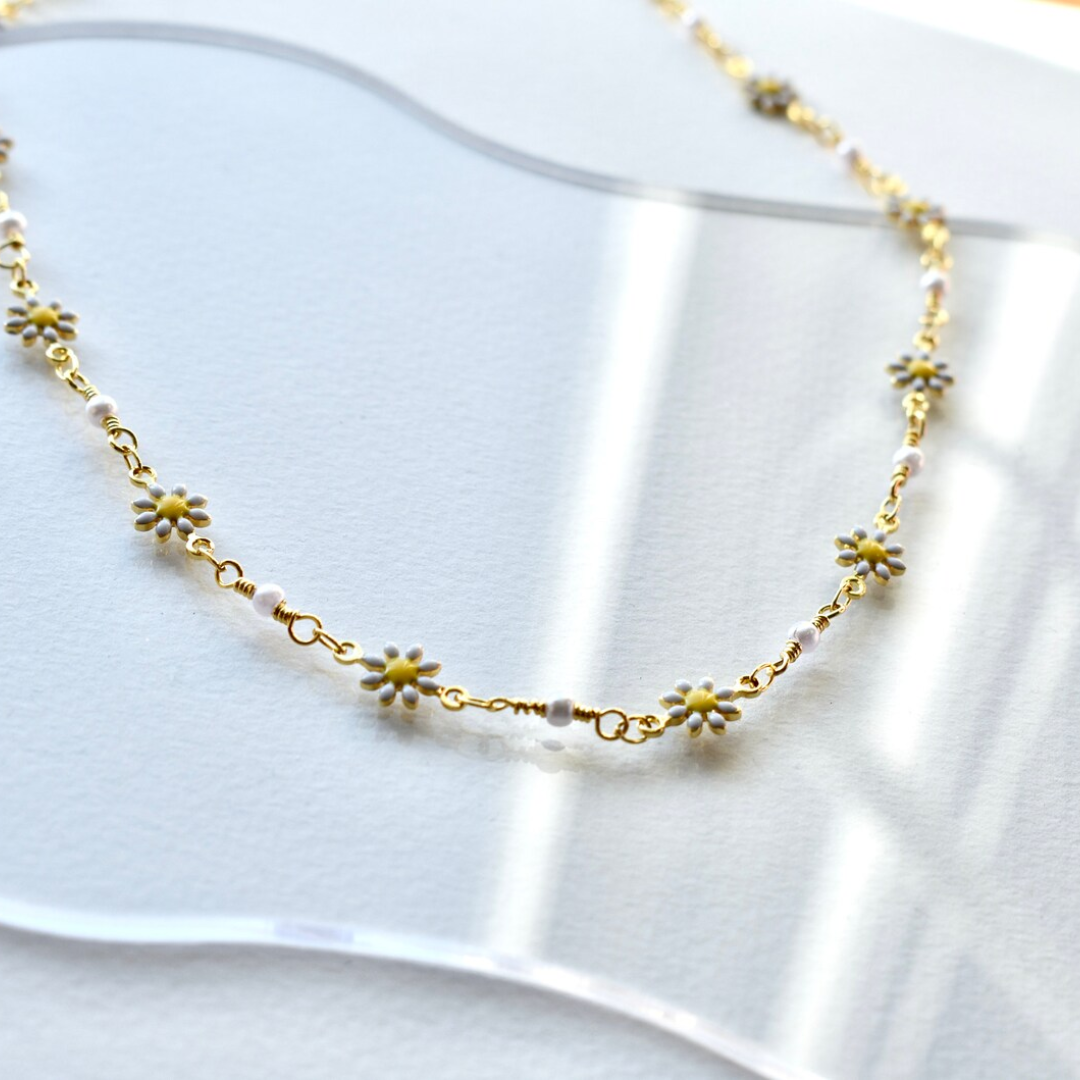 A delicate gold chain necklace adorned with small white daisies. The daisies have yellow centers and are evenly spaced along the chain.