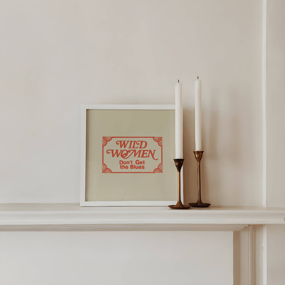 Framed artwork with 'Wild Women Don't Get the Blues' text on a shelf with two candles.