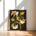 A framed art print featuring a retro floral design with cream flowers, yellow-green leaves, and small white stars on a dark charcoal background, leaning against a wall on a light wood floor.