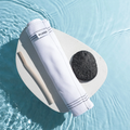 A white, textured exfoliating towel is rolled up and placed on a round, smooth surface. Next to it is a black loofah and a wooden stick. The background is a pool of blue water.
