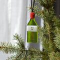 A small green bottle-shaped ornament with a white band, simple smiley face, and red cap hanging from a black string on a pine tree branch.