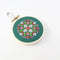 A small, circular embroidery hoop with a green background. The hoop features a colorful cross-stitch pattern with pink, white, and red geometric shapes. There is a metal ring attached to the hoop for hanging.