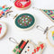 A small, circular embroidery hoop with a green background. The hoop features a colorful cross-stitch pattern with pink, white, and red geometric shapes. There is a metal ring attached to the hoop for hanging.