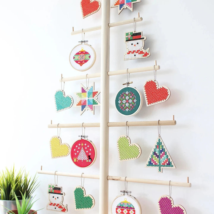 A small, circular embroidery hoop with a green background. The hoop features a colorful cross-stitch pattern with pink, white, and red geometric shapes. There is a metal ring attached to the hoop for hanging.