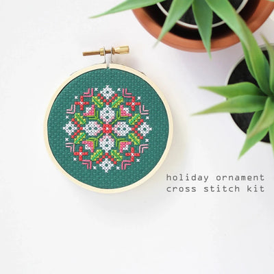 A small, circular embroidery hoop with a green background. The hoop features a colorful cross-stitch pattern with pink, white, and red geometric shapes. There is a metal ring attached to the hoop for hanging.