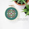 A small, circular embroidery hoop with a green background. The hoop features a colorful cross-stitch pattern with pink, white, and red geometric shapes. There is a metal ring attached to the hoop for hanging.