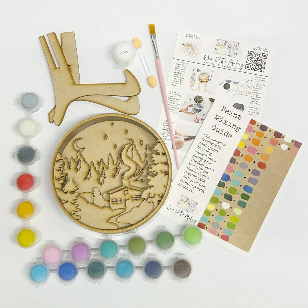 A Paint By Layers Craft Kit featuring a circular shadow box with a pre-designed winter scene. The scene depicts a cabin in a snowy forest under a starry night sky. The kit includes layers of wood, paint, and instructions. The title of the kit is "Winter Cabin Shadow Box."