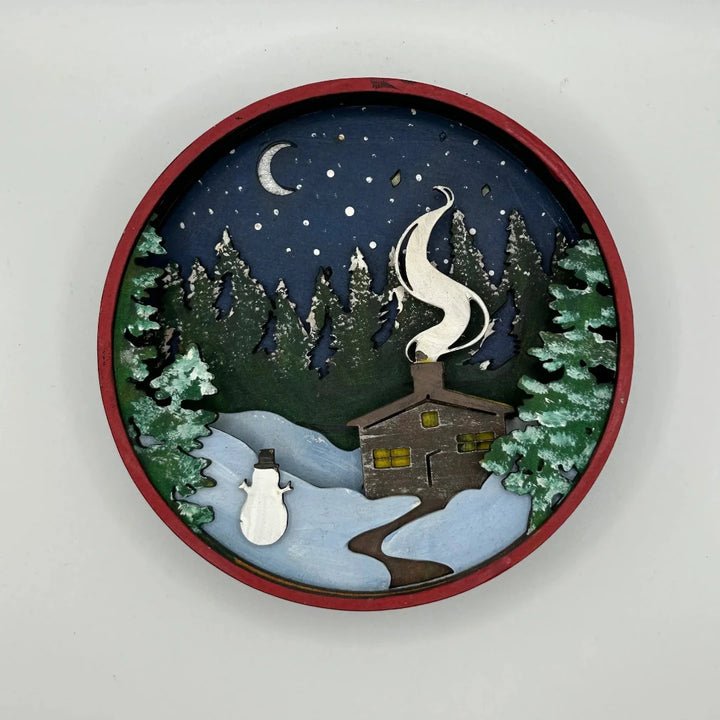 A Paint By Layers Craft Kit featuring a circular shadow box with a pre-designed winter scene. The scene depicts a cabin in a snowy forest under a starry night sky. The kit includes layers of wood, paint, and instructions. The title of the kit is 