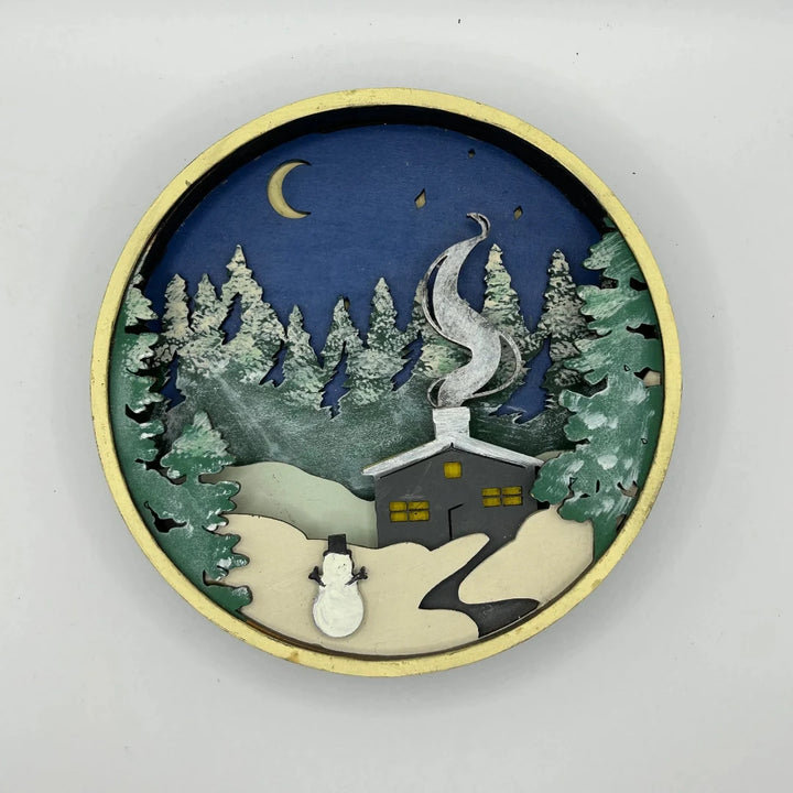 A Paint By Layers Craft Kit featuring a circular shadow box with a pre-designed winter scene. The scene depicts a cabin in a snowy forest under a starry night sky. The kit includes layers of wood, paint, and instructions. The title of the kit is 