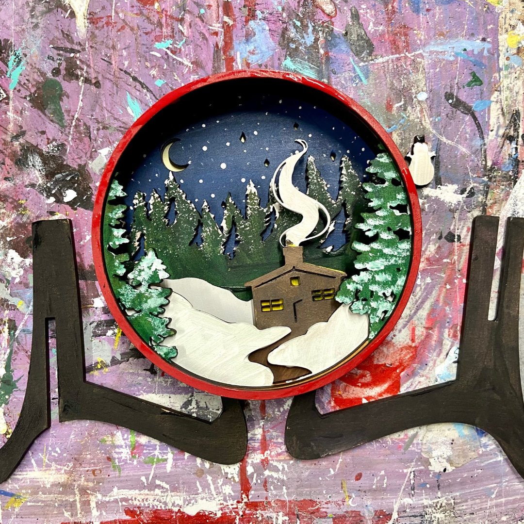 A Paint By Layers Craft Kit featuring a circular shadow box with a pre-designed winter scene. The scene depicts a cabin in a snowy forest under a starry night sky. The kit includes layers of wood, paint, and instructions. The title of the kit is "Winter Cabin Shadow Box."