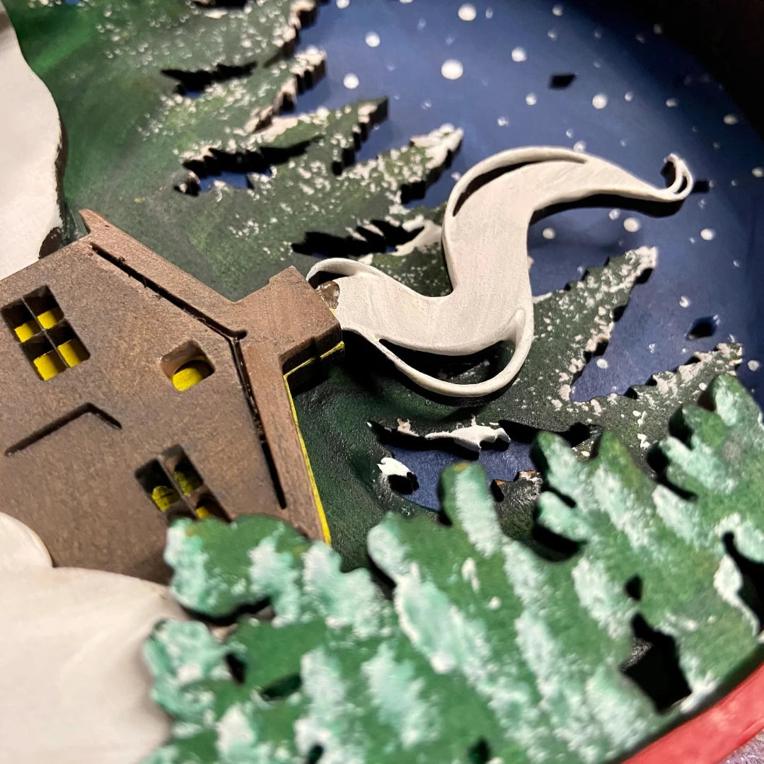 A Paint By Layers Craft Kit featuring a circular shadow box with a pre-designed winter scene. The scene depicts a cabin in a snowy forest under a starry night sky. The kit includes layers of wood, paint, and instructions. The title of the kit is "Winter Cabin Shadow Box."