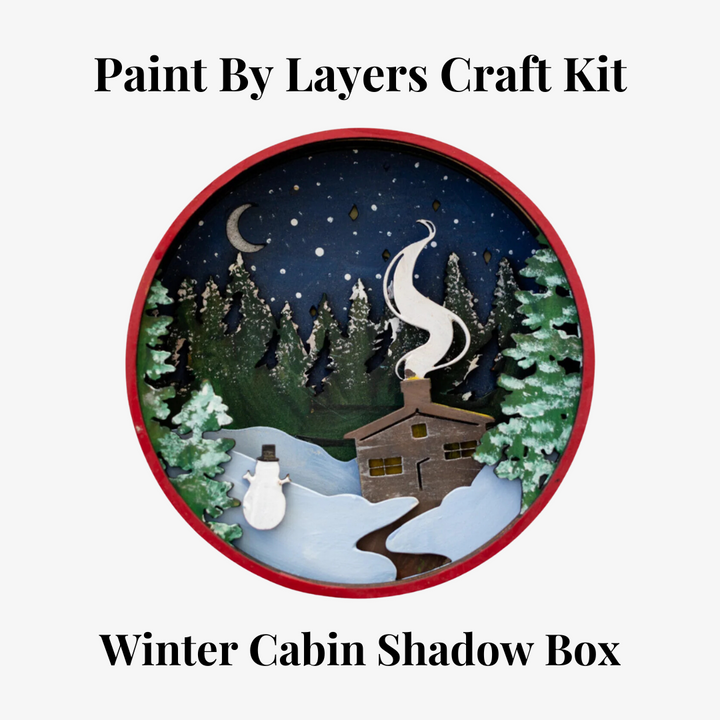 A Paint By Layers Craft Kit featuring a circular shadow box with a pre-designed winter scene. The scene depicts a cabin in a snowy forest under a starry night sky. The kit includes layers of wood, paint, and instructions. The title of the kit is 